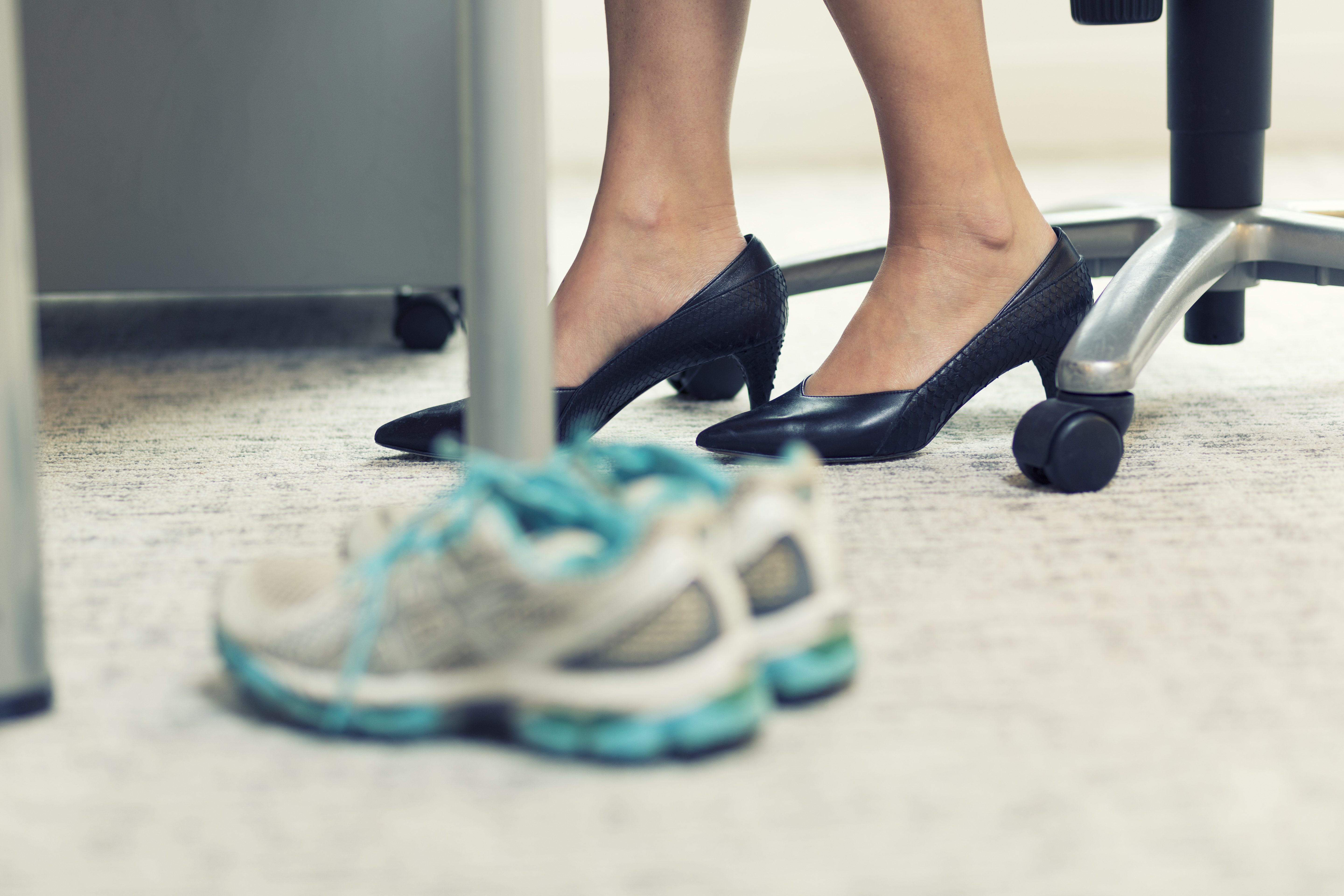 Women's feet in pumps under a desk and safety shoes in the foreground.