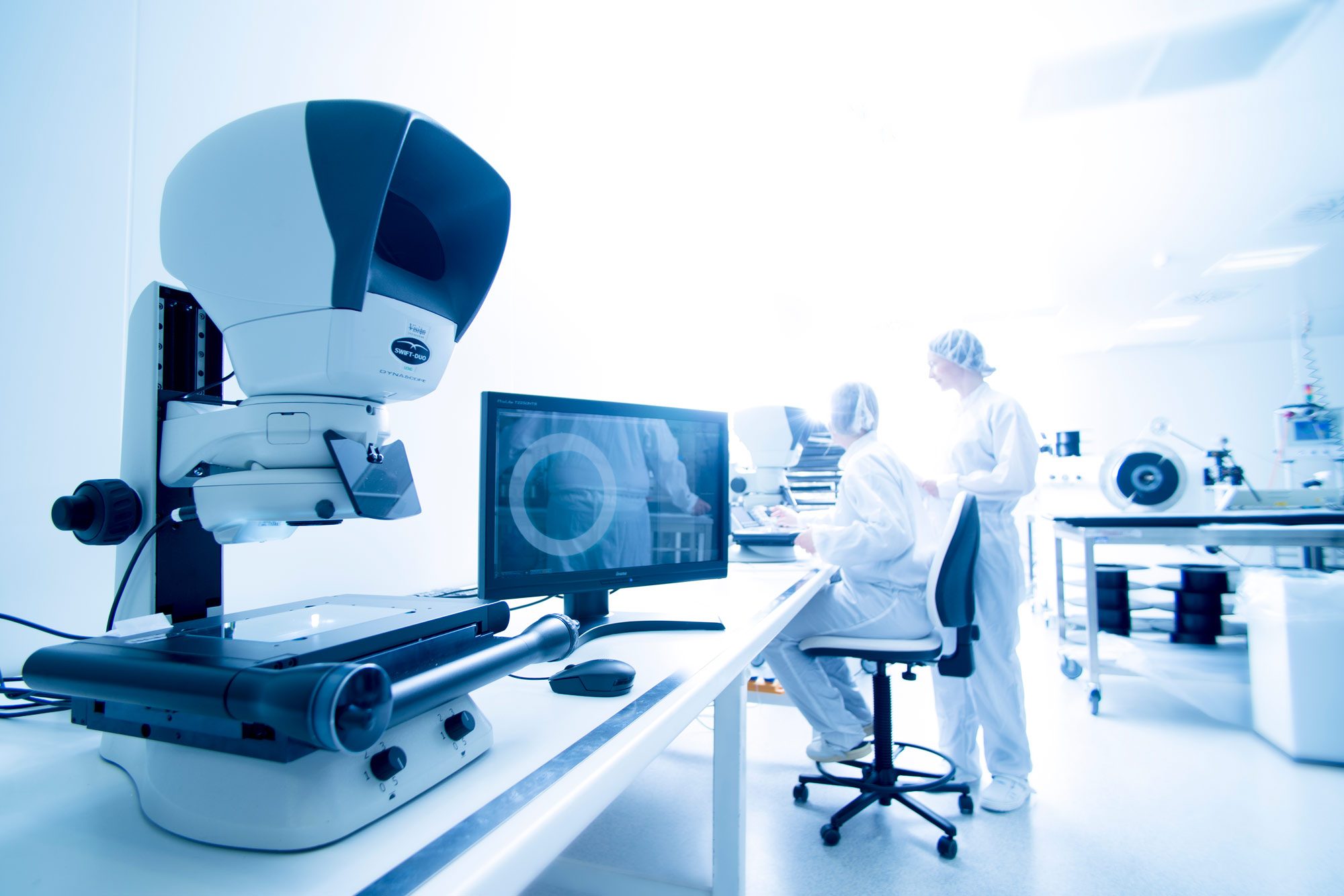 Two employees work in the clean room and a microscope and a screen are in the foreground. 